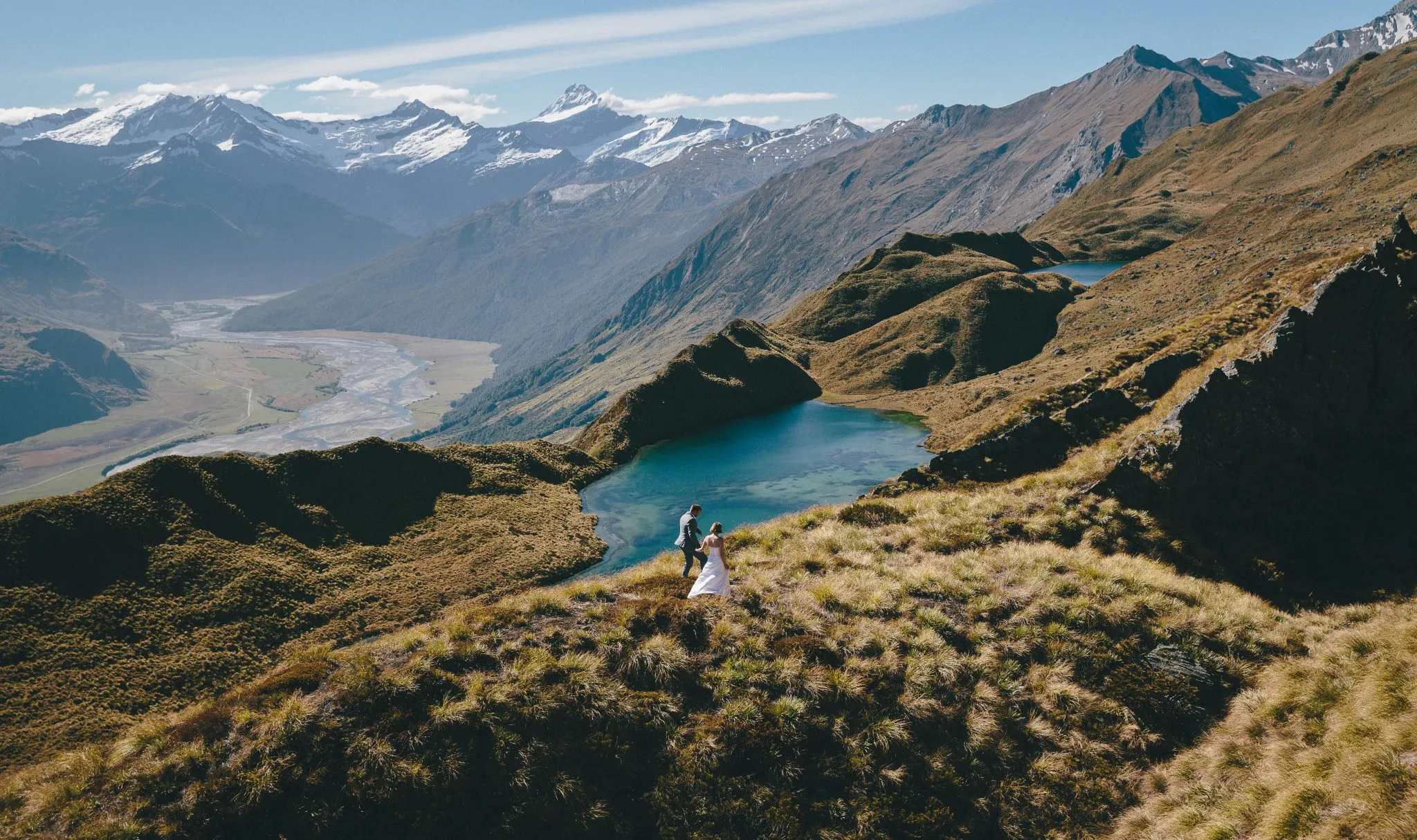 A bride and groom walking above a crystal-clear alpine tarn at Alta Tarns, with Tititea / Mount Aspiring beyond