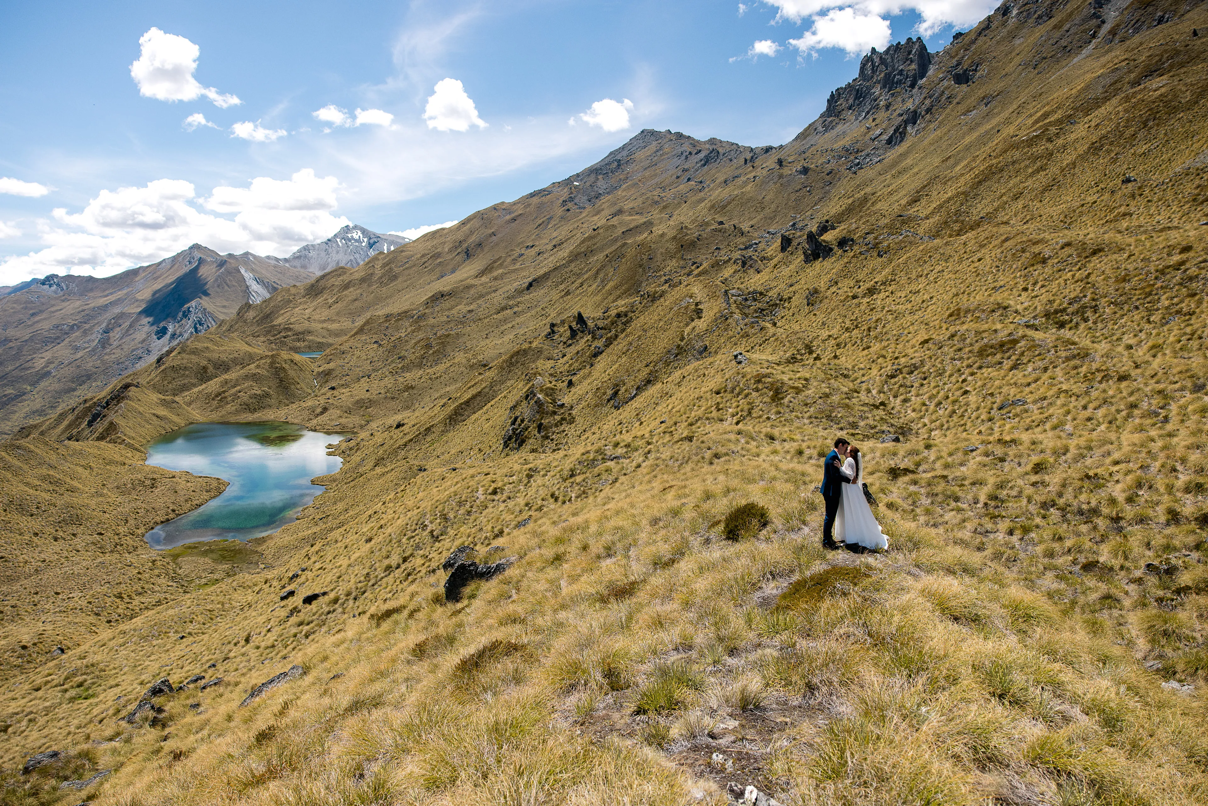 A couple embracing on the golden tussock above a mountain tarn at Alta Tarns