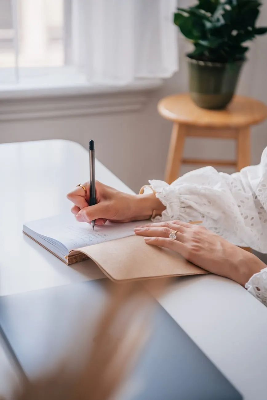 Woman writing by hand in a notebook at a desk by a window