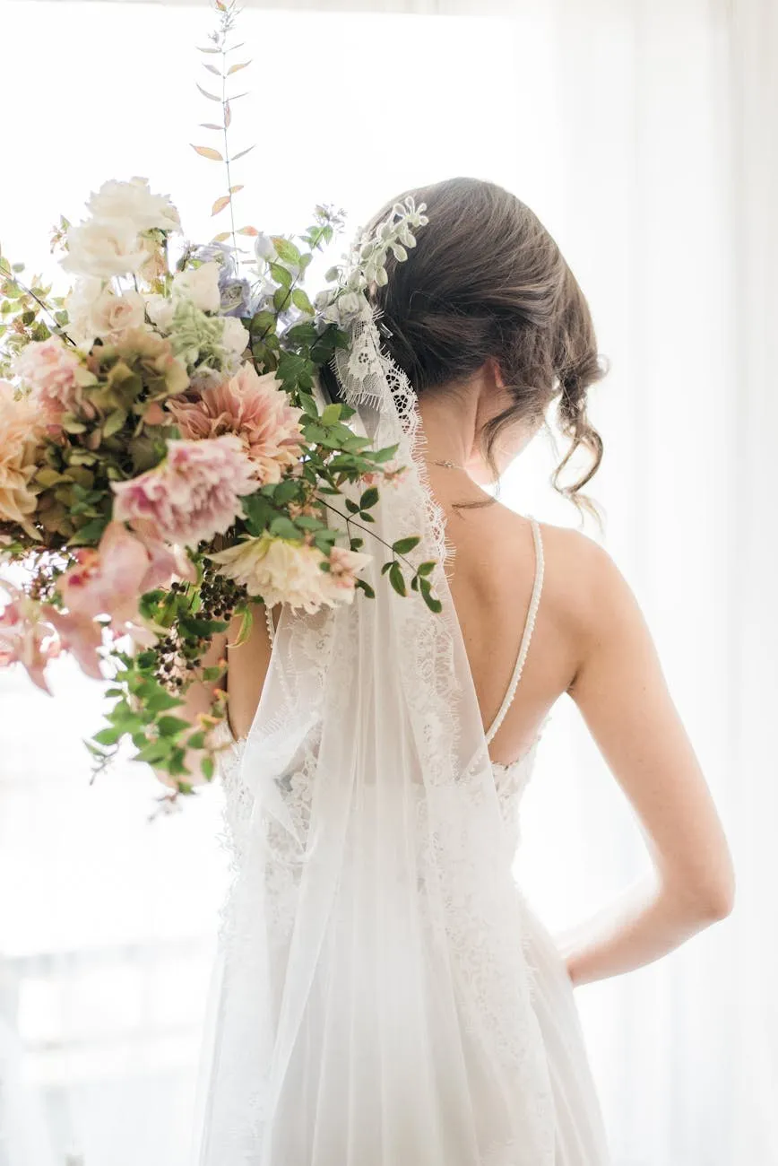 Bride seen from behind holding a large bouquet beside a bright window