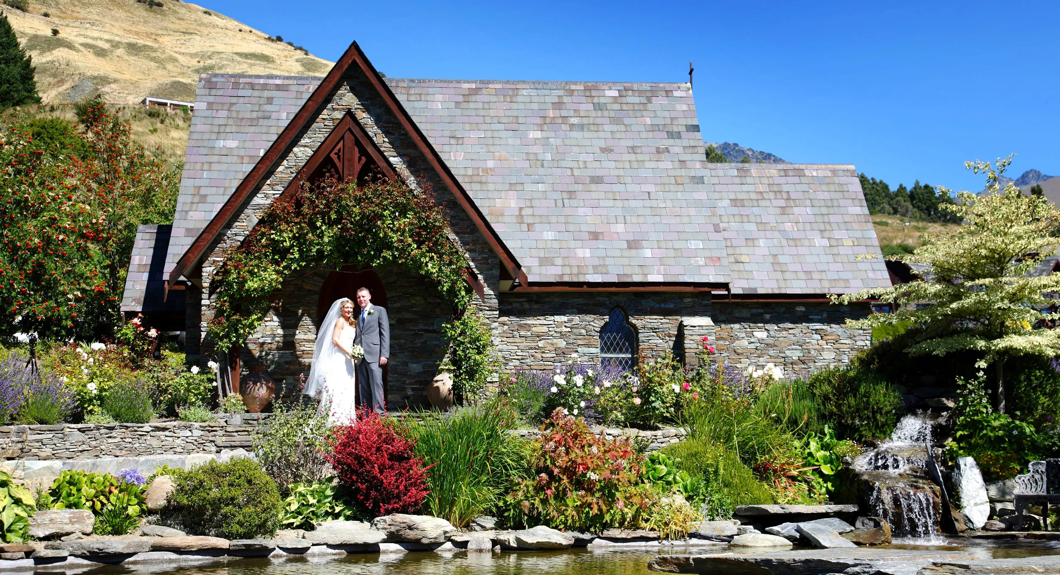 A bride and groom in front of the stone chapel and gardens at Stoneridge Estate, Queenstown