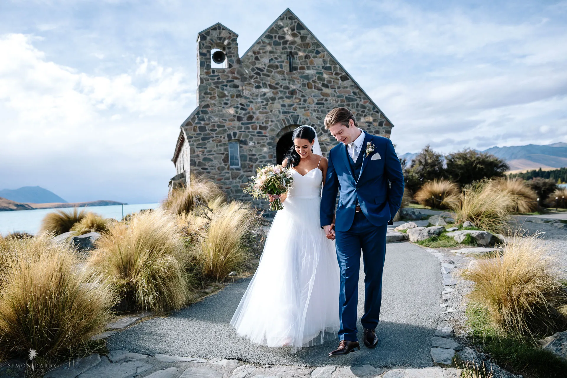 A newly married couple walk hand in hand past the stone Church of the Good Shepherd on the shore of Lake Tekapo