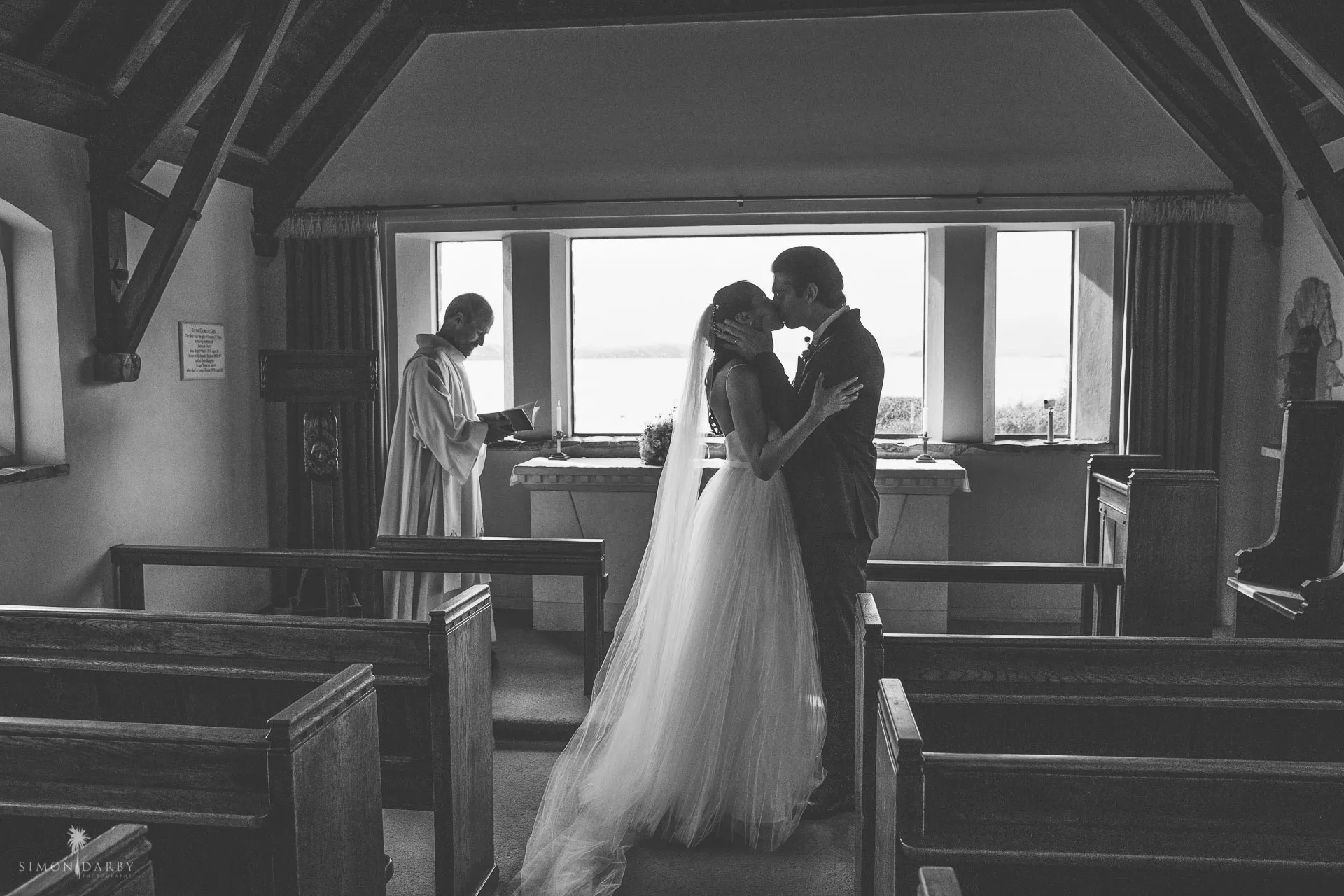 A couple share their first kiss during the ceremony inside the Church of the Good Shepherd, the minister and lake window behind them