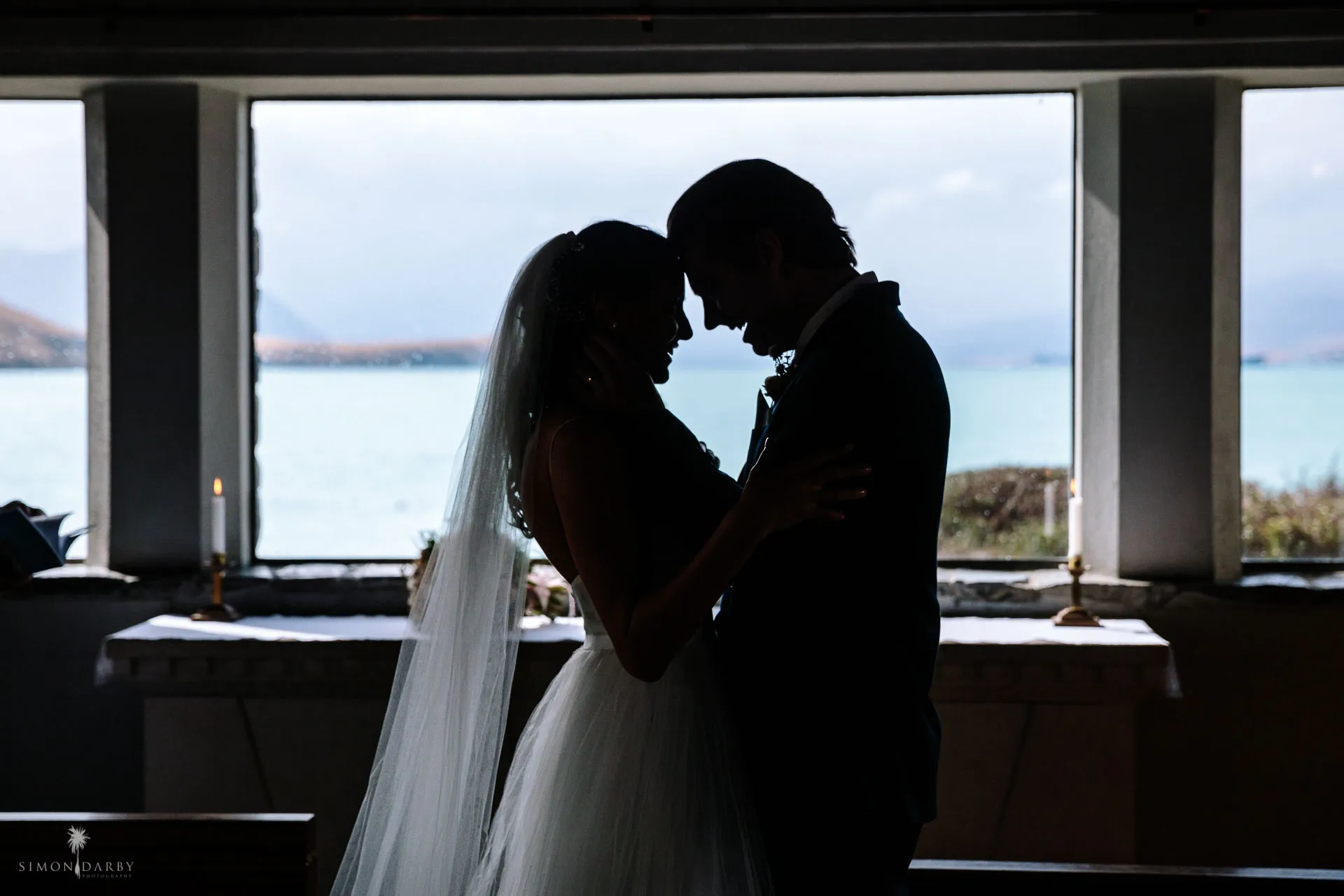 A bride and groom silhouetted against the altar window overlooking Lake Tekapo inside the chapel