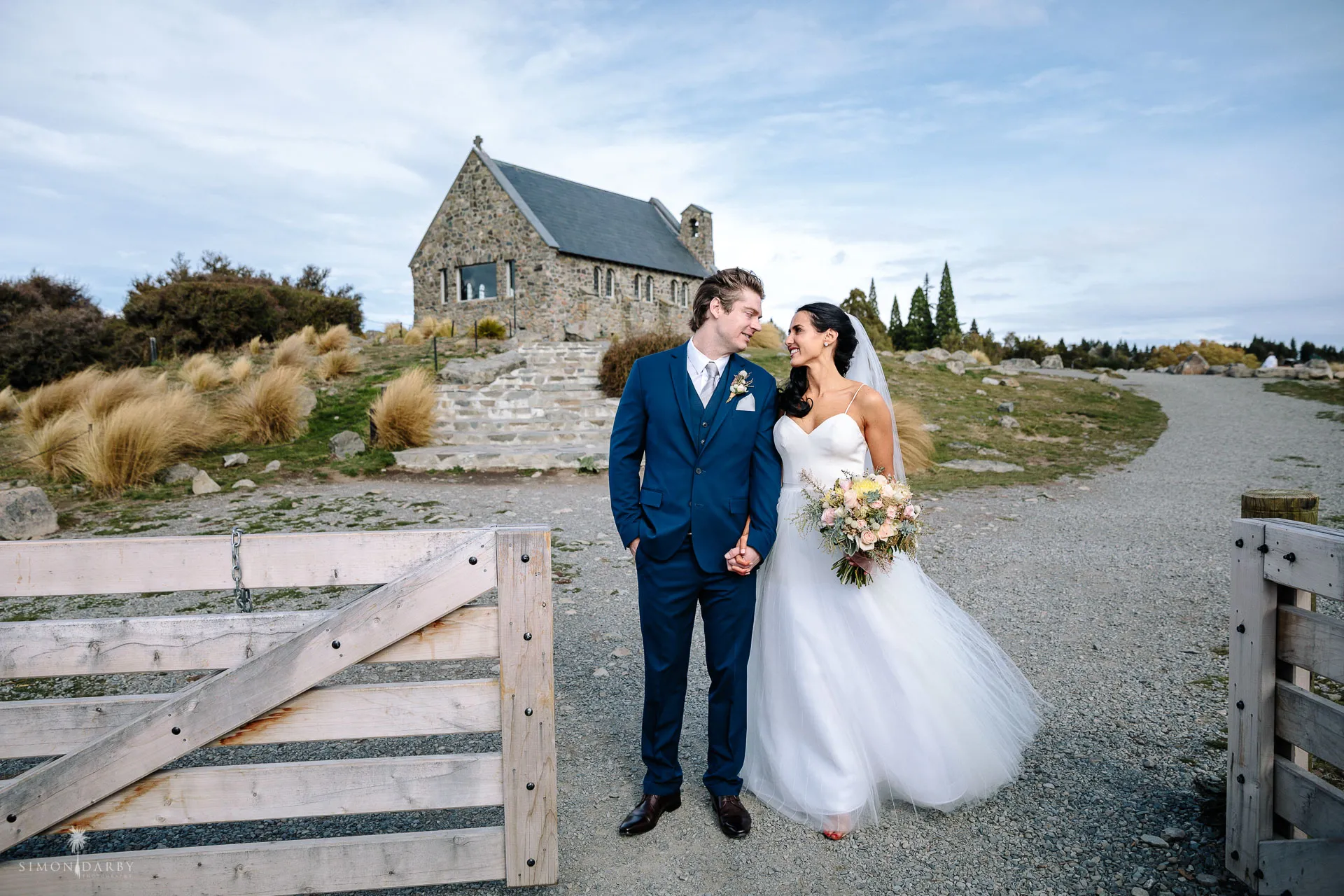 A couple pause for a portrait on the tussock hillside with the Church of the Good Shepherd behind them