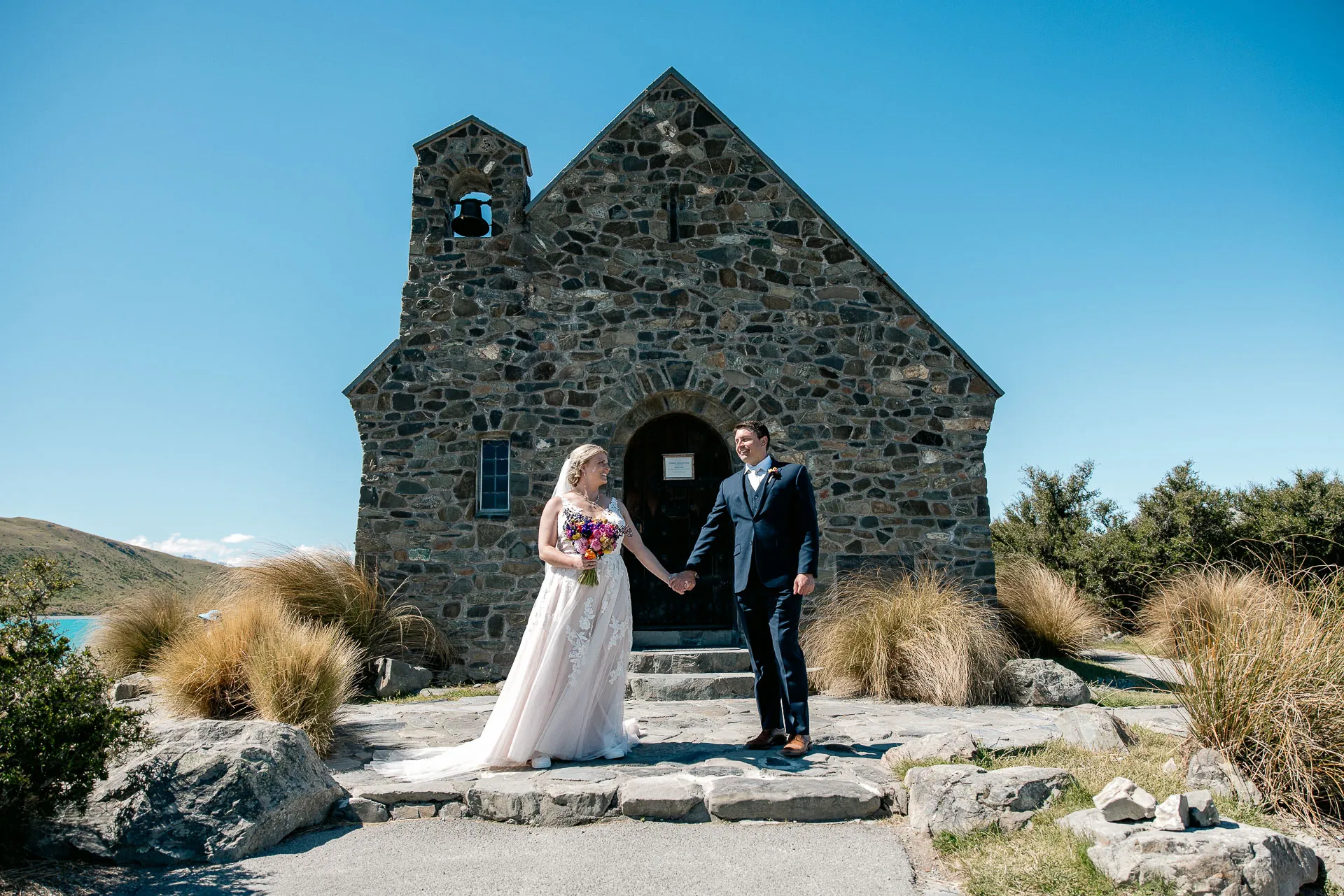 A bride and groom outside the front of the historic stone Church of the Good Shepherd under a clear blue Tekapo sky