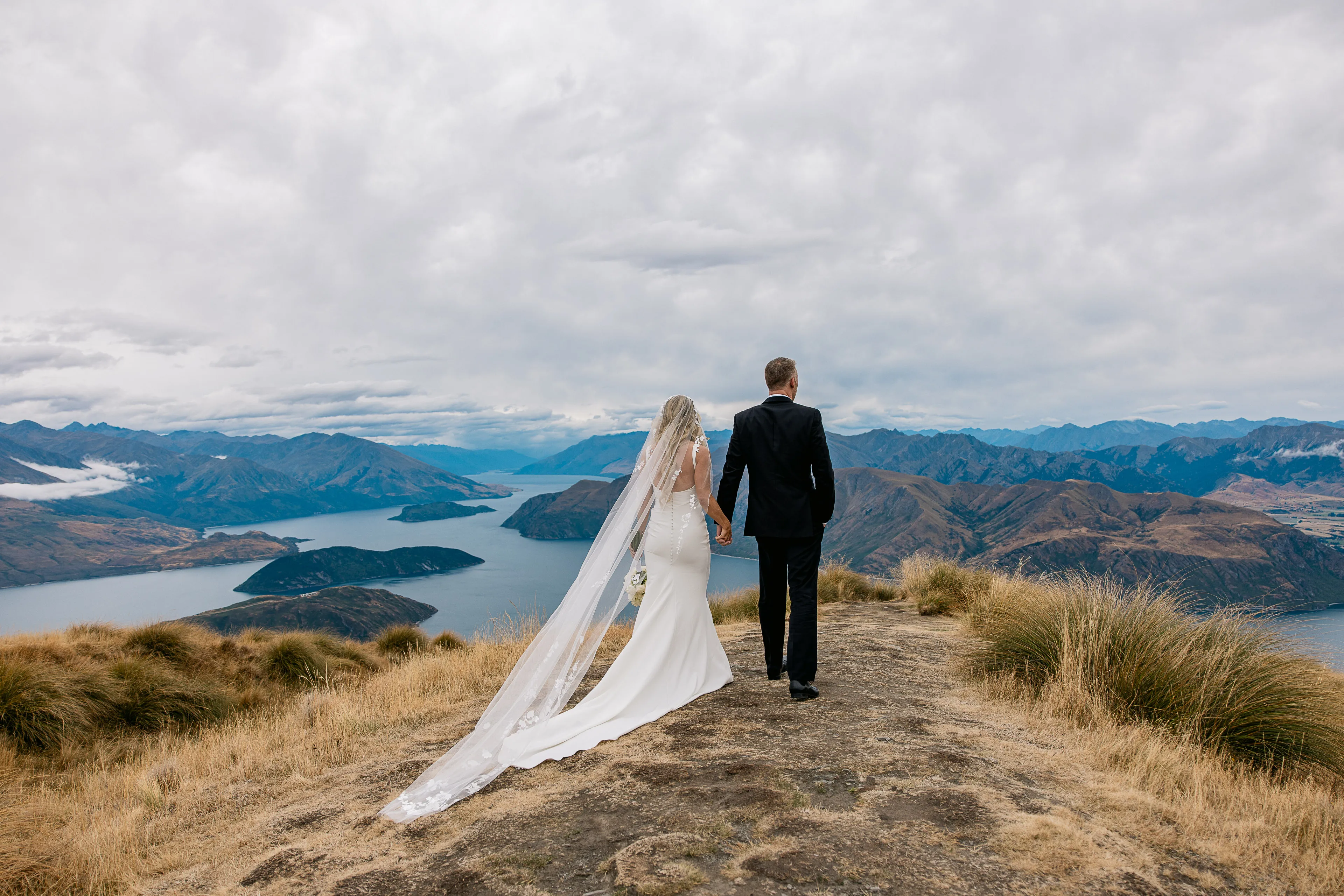 A bride and groom walking hand in hand along the Coromandel Peak ridgeline above Lake Wānaka
