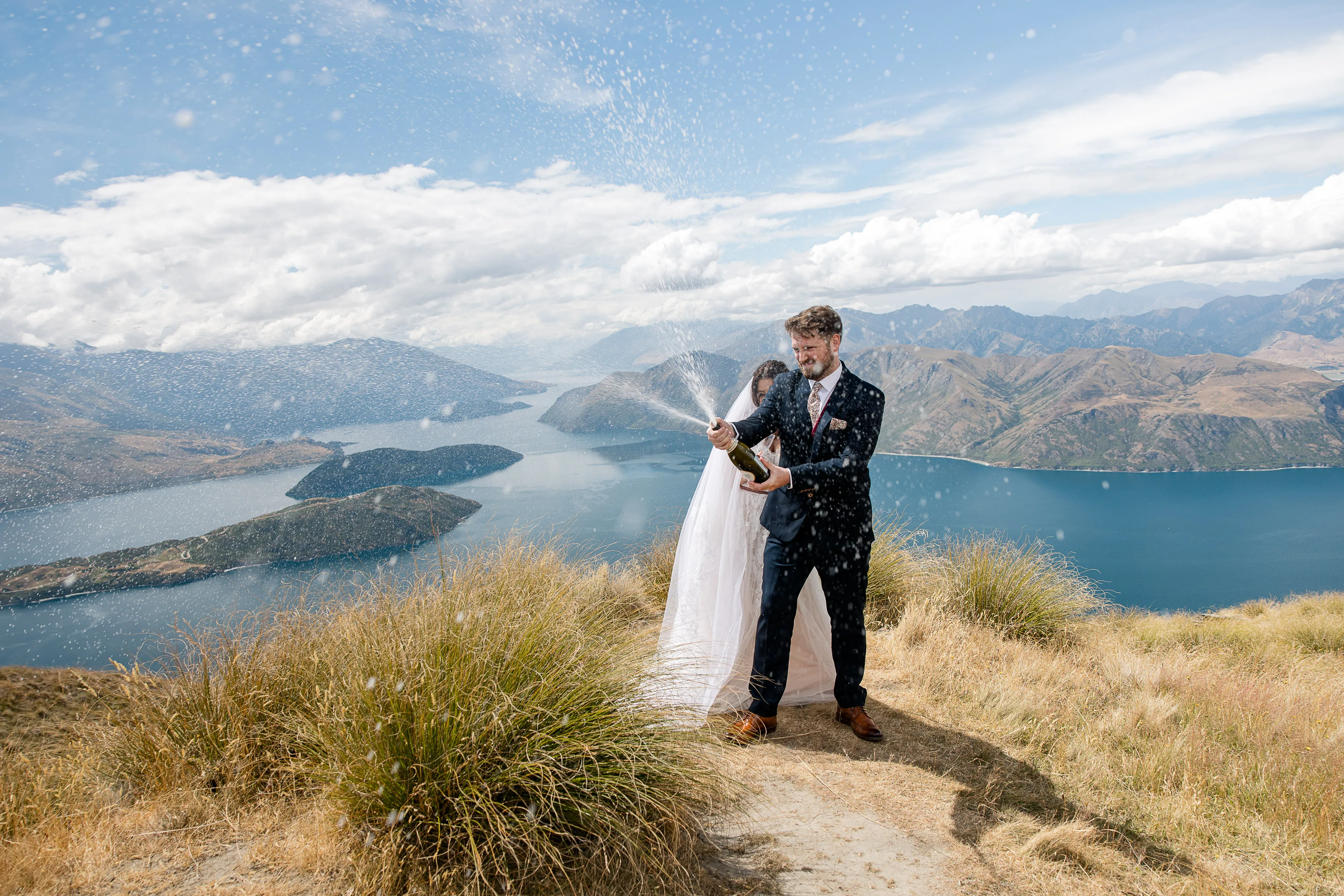 A bride and groom celebrating with champagne on Coromandel Peak above Lake Wānaka