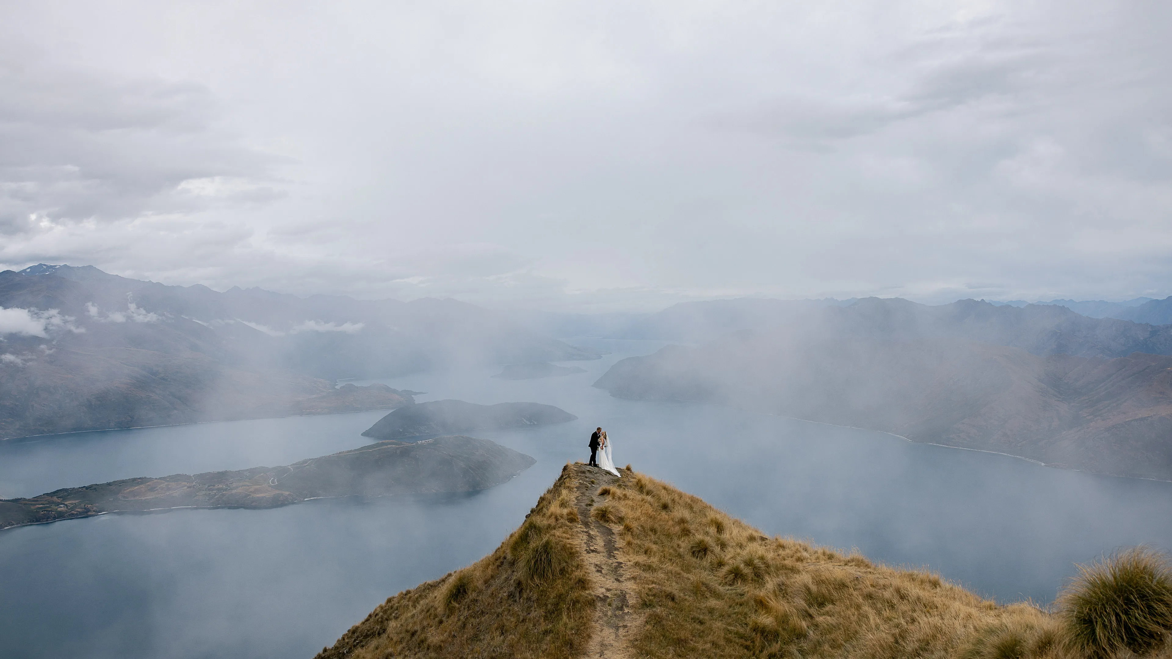 A couple embracing on Coromandel Peak as cloud drifts over Lake Wānaka below