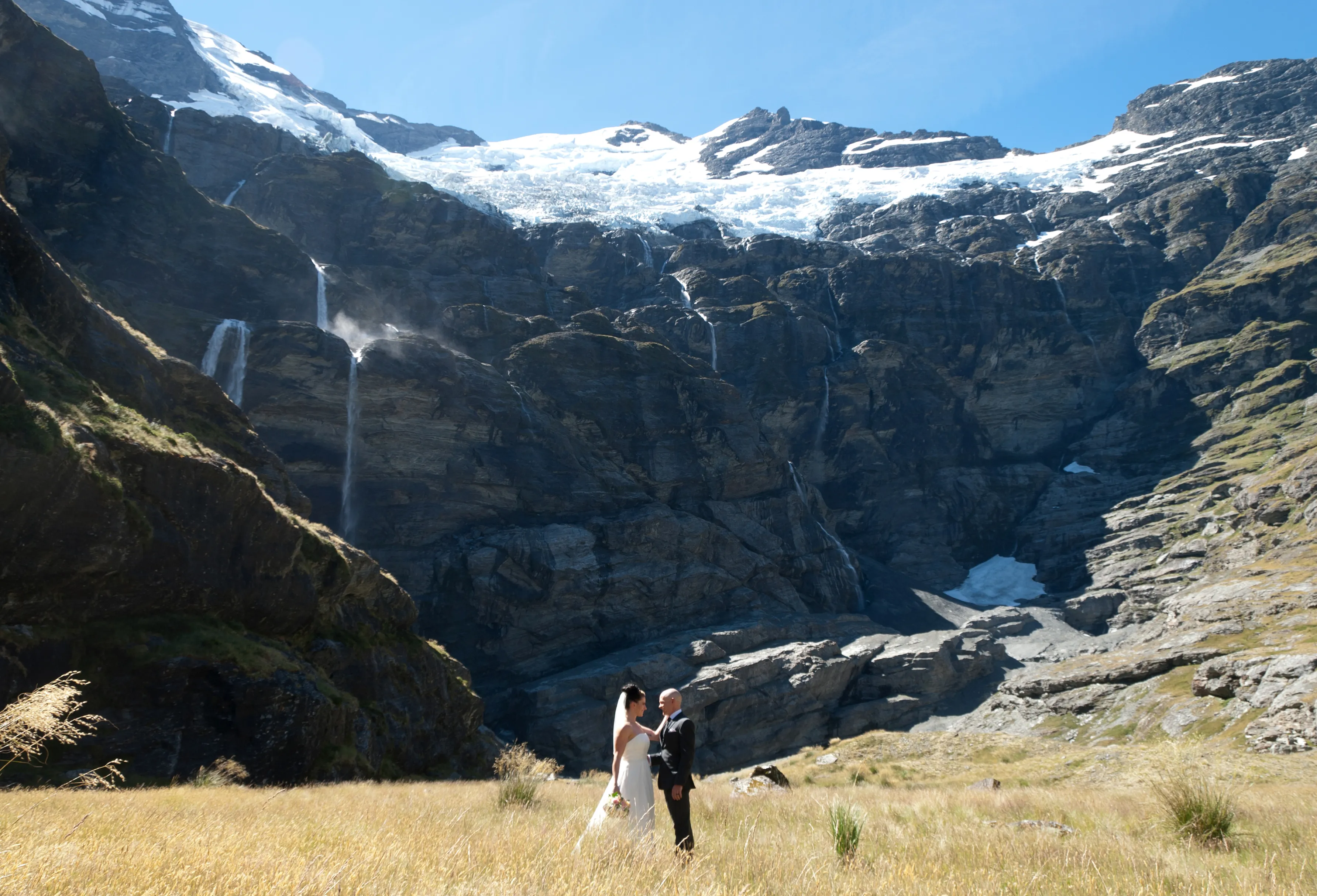 A couple embracing in the golden tussock of Earnslaw Burn, waterfalls tumbling behind them