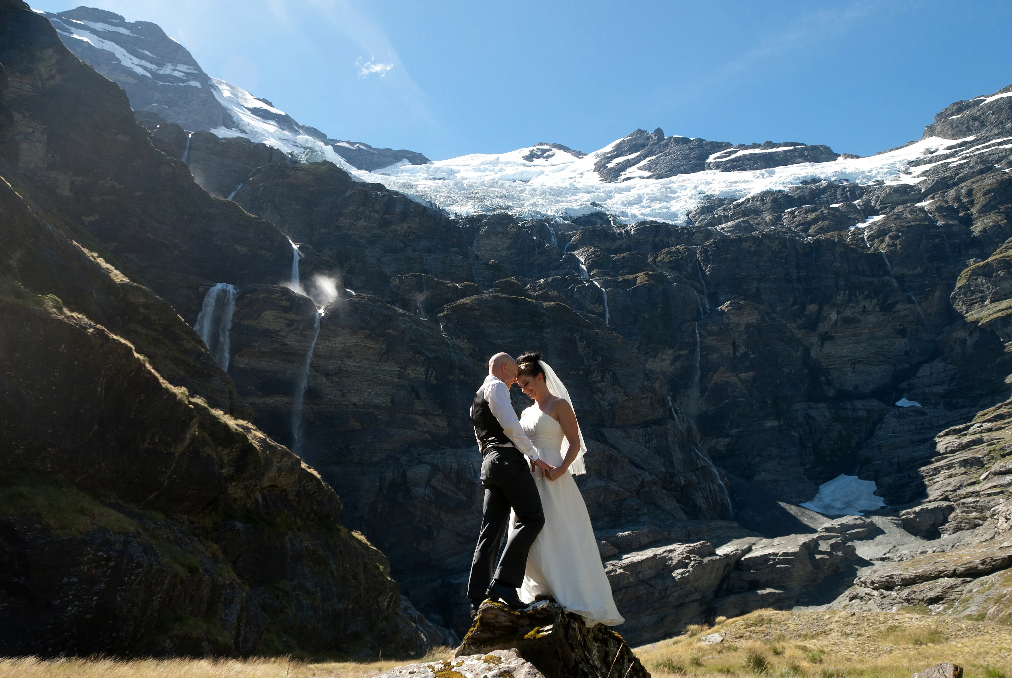 A couple embracing on a rock at Earnslaw Burn beneath a hanging glacier and waterfalls
