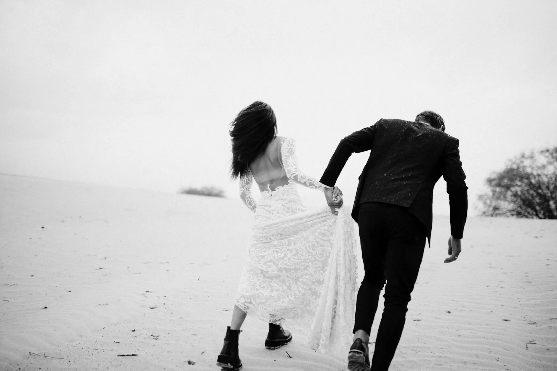 Bride and groom walking hand in hand across sand dunes, black and white