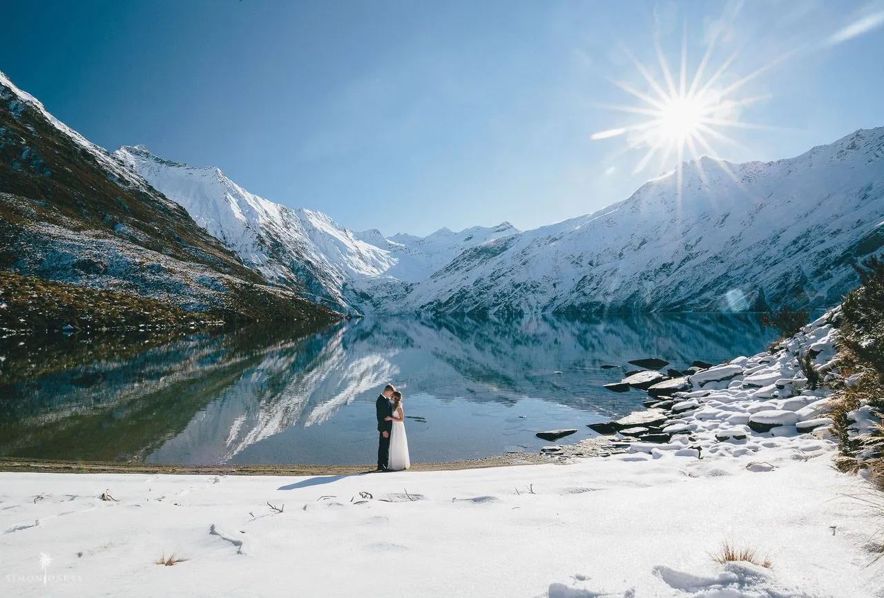 A couple embracing on the snowy shore of Lake Lochnagar, snow-capped peaks reflected in the still alpine lake