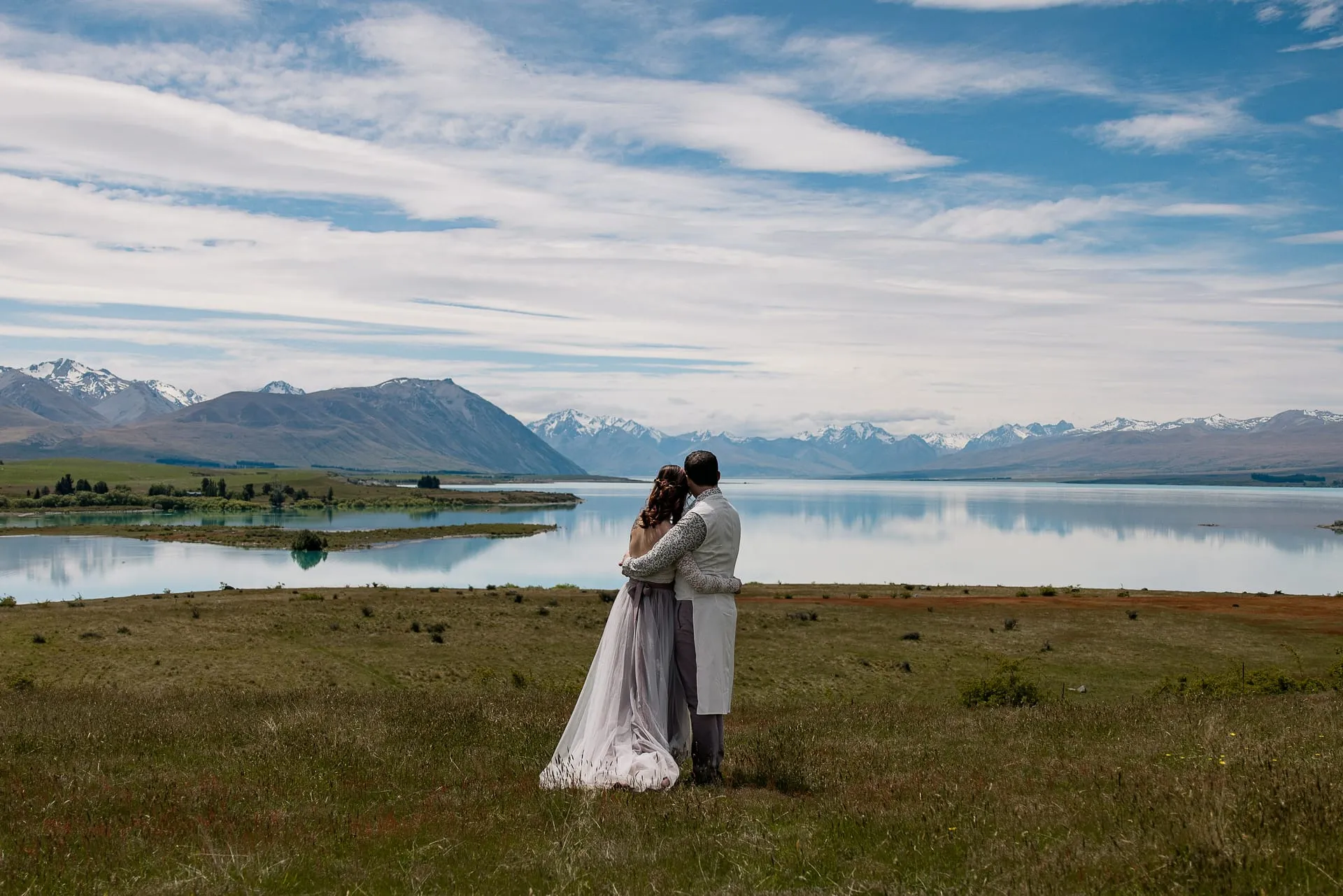 A couple embracing on a hilltop above turquoise Lake Tekapo, with snow-capped mountains beyond