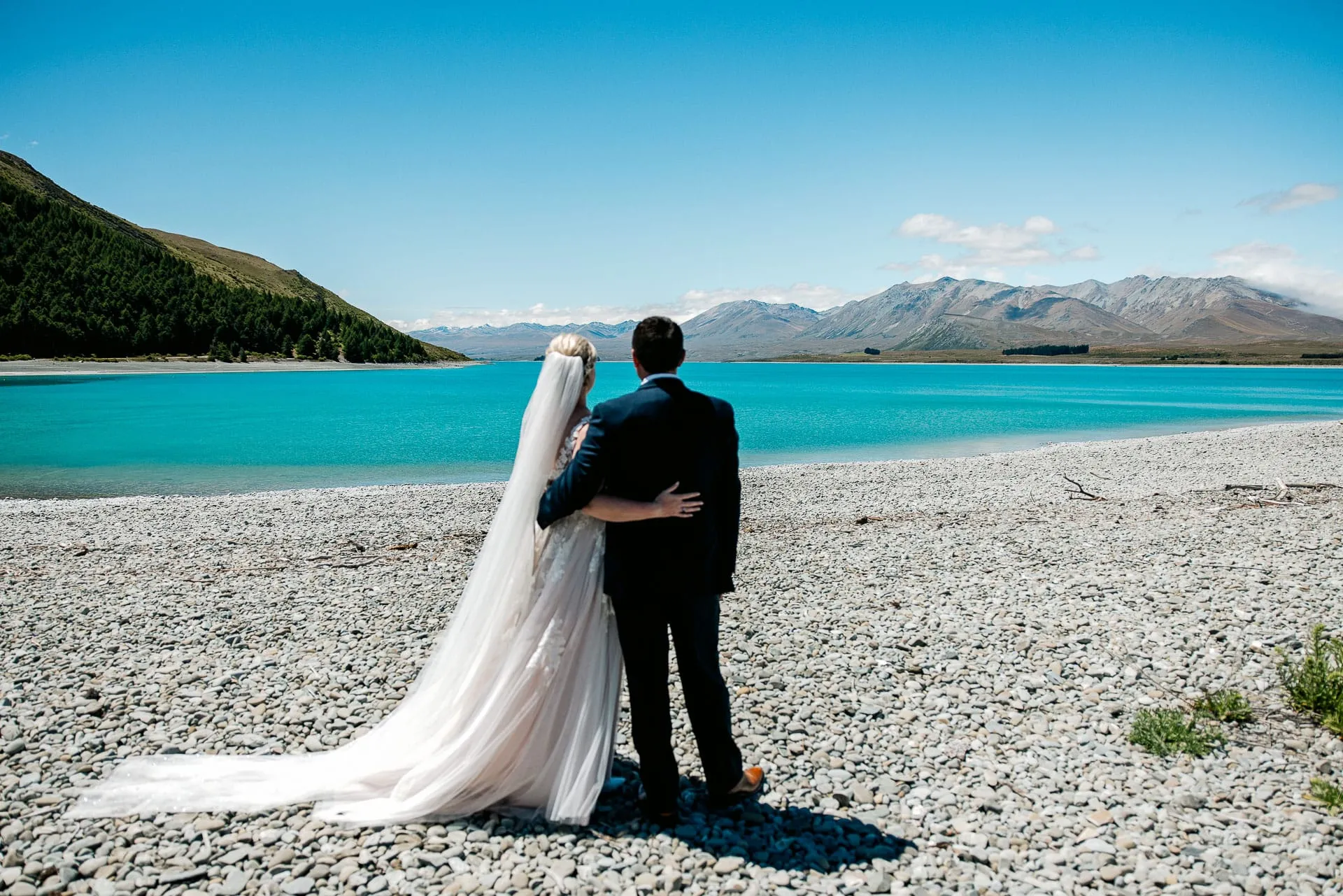 A bride and groom on the pebble shore of turquoise Lake Tekapo, looking out to the mountains