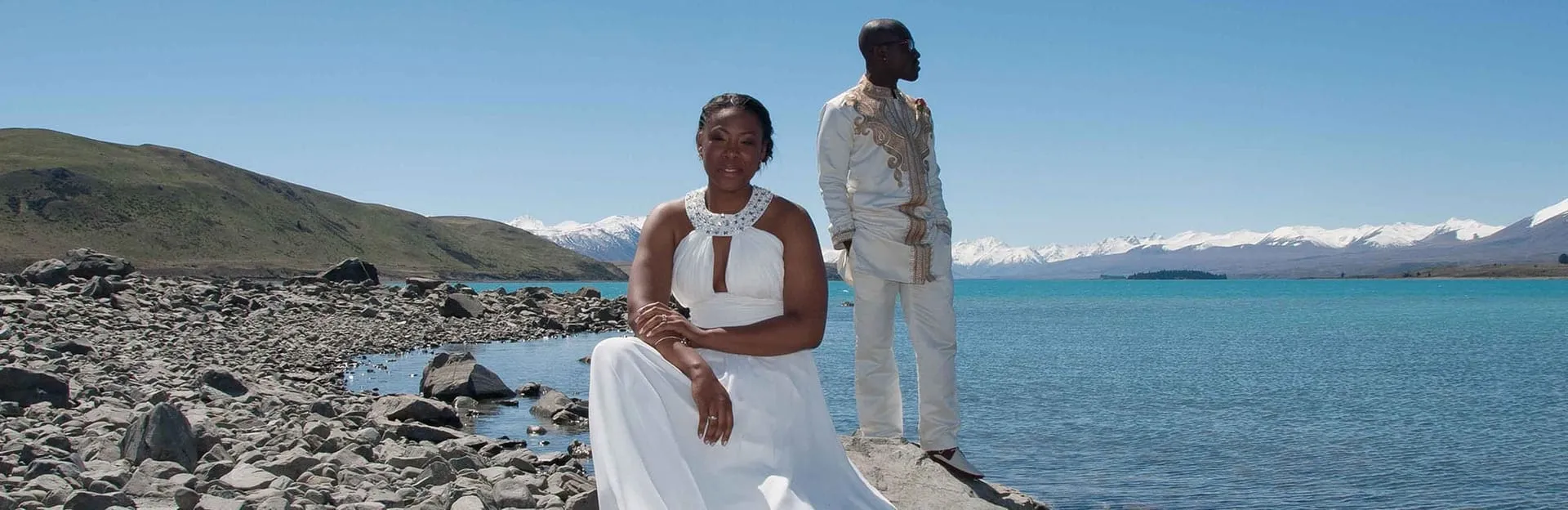 A couple in white and gold attire on the rocky shore of Lake Tekapo, snow-capped mountains behind