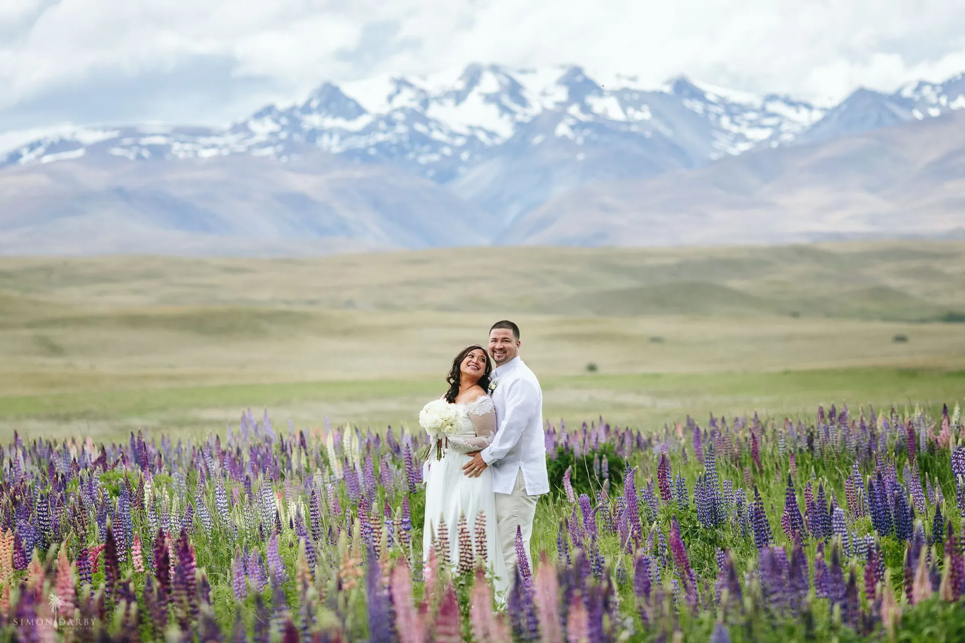 Newlyweds embracing in a field of lupins with snow-capped mountains behind at Lake Tekapo