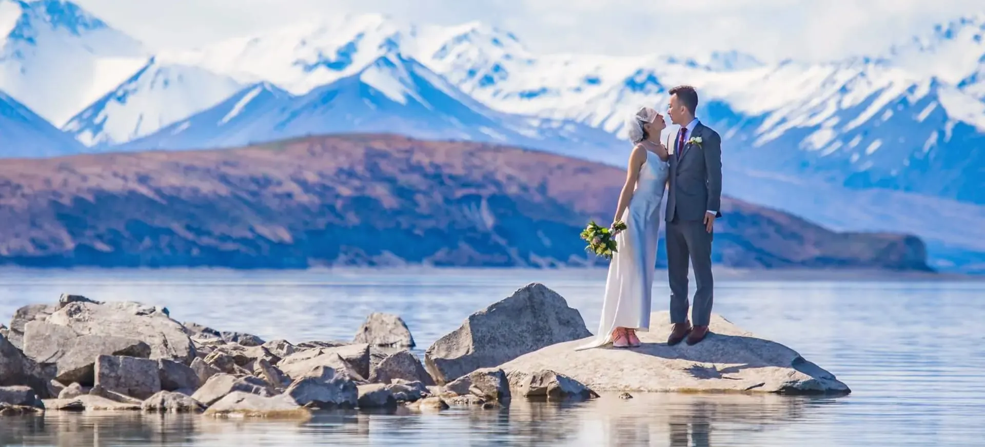 A couple sharing a kiss on a rock in Lake Tekapo, beneath the snow-capped Southern Alps