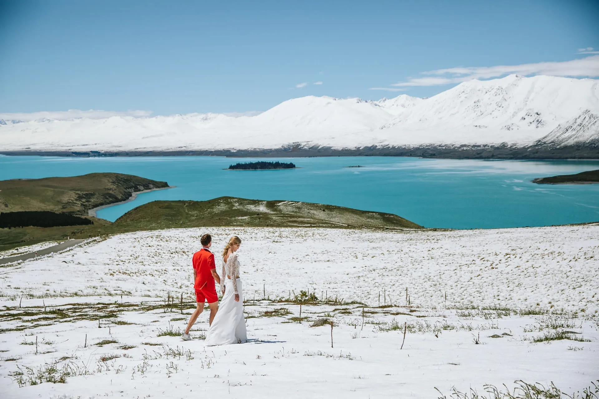 A couple walking a snow-covered hilltop above turquoise Lake Tekapo, with snow-capped mountains behind