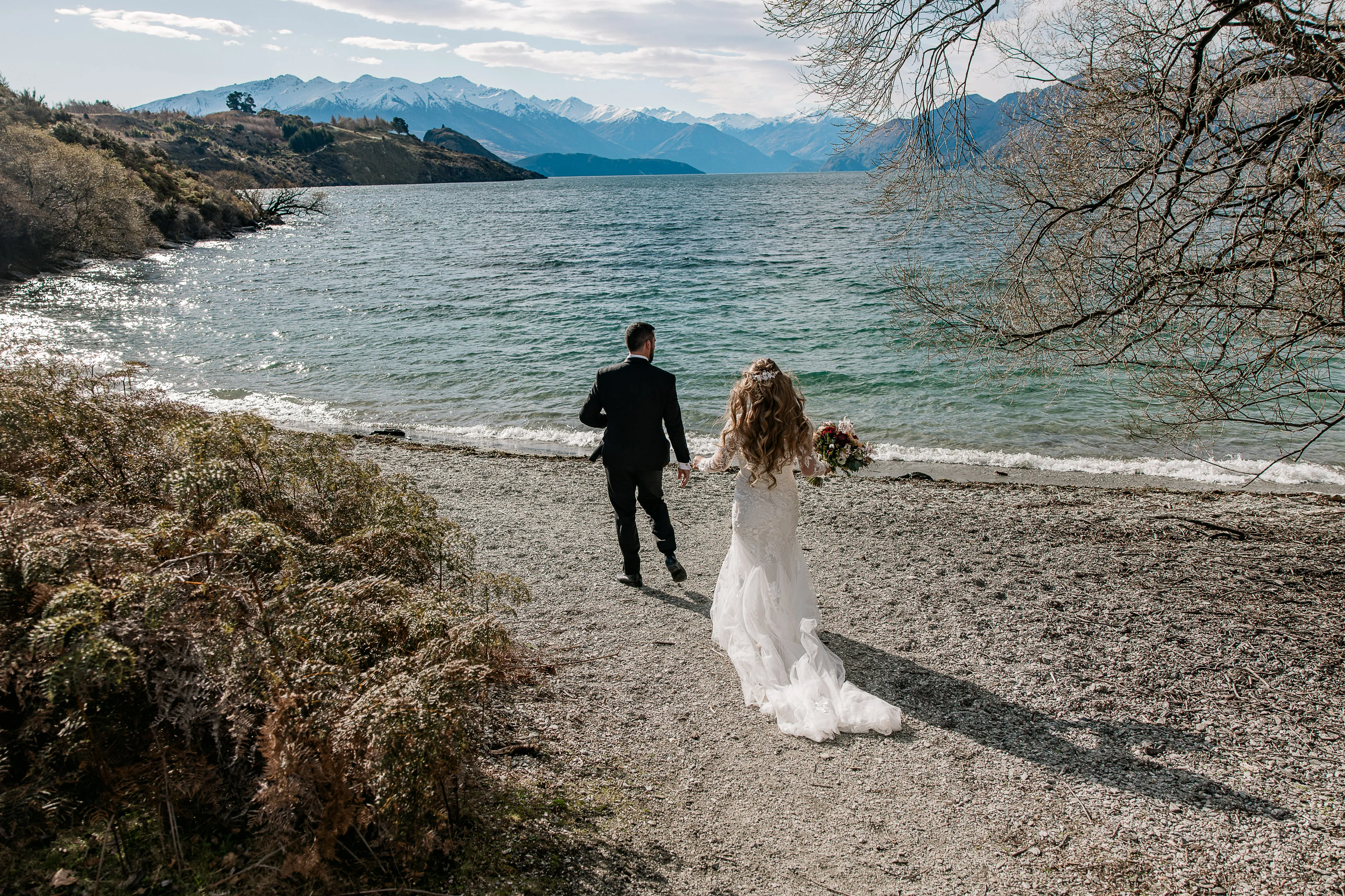 A bride and groom walk hand in hand along the pebble shore of Lake Wānaka, snow-capped mountains across the water
