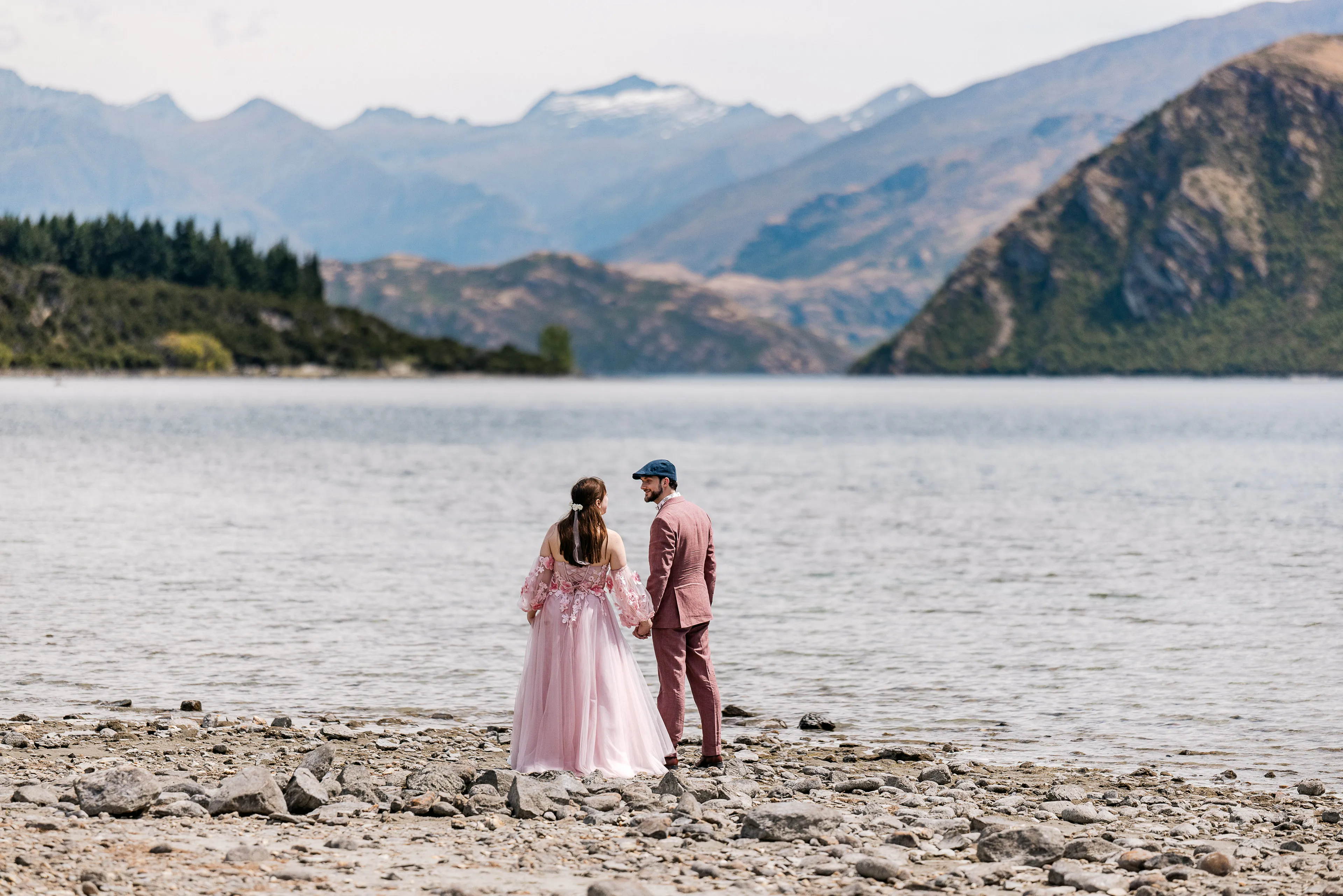 A bride in a blush gown and groom in a dusty-pink suit stand hand in hand on the rocky Lake Wānaka shore
