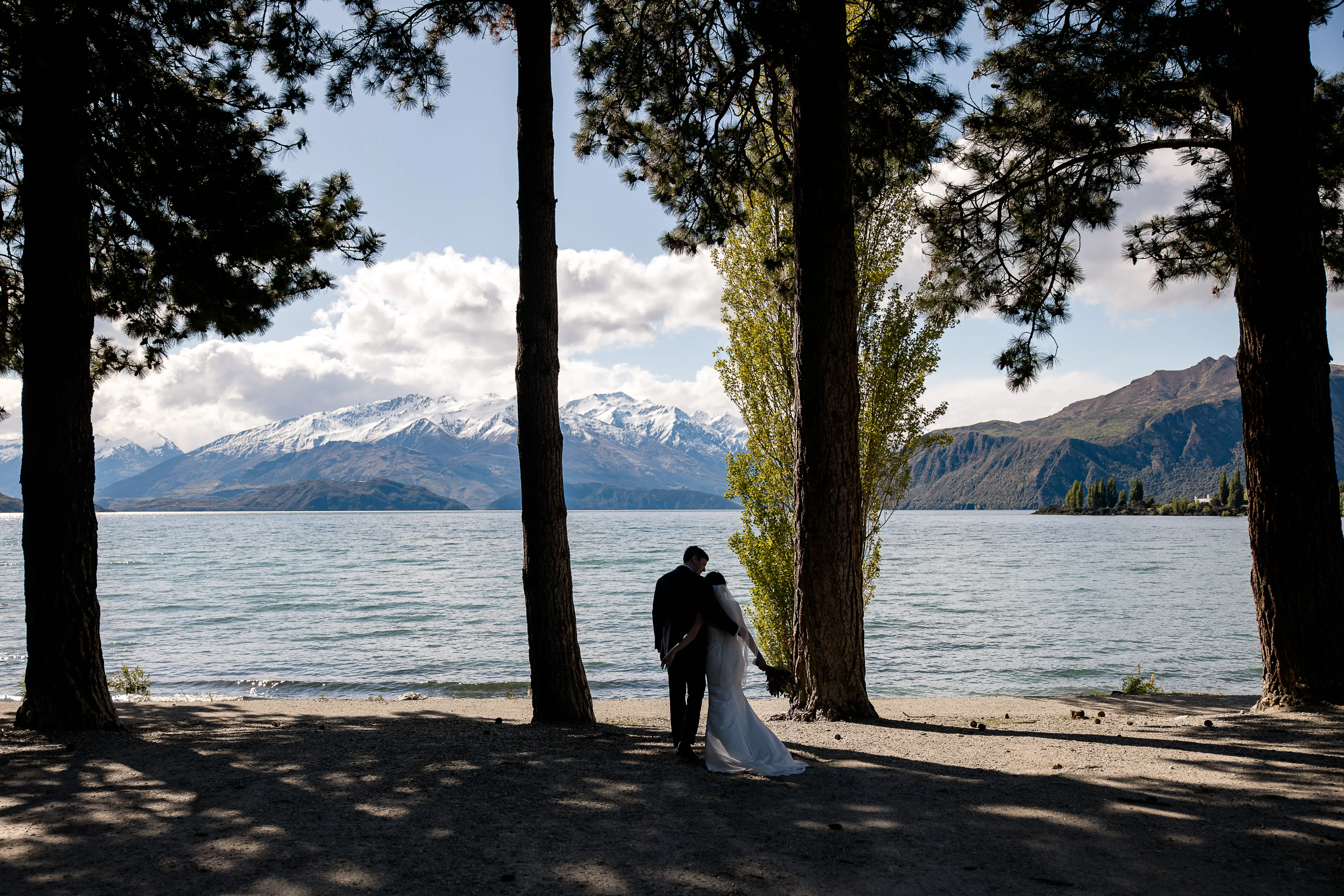 A couple embrace among tall trees on the shore of Lake Wānaka, snow-capped mountains framed beyond