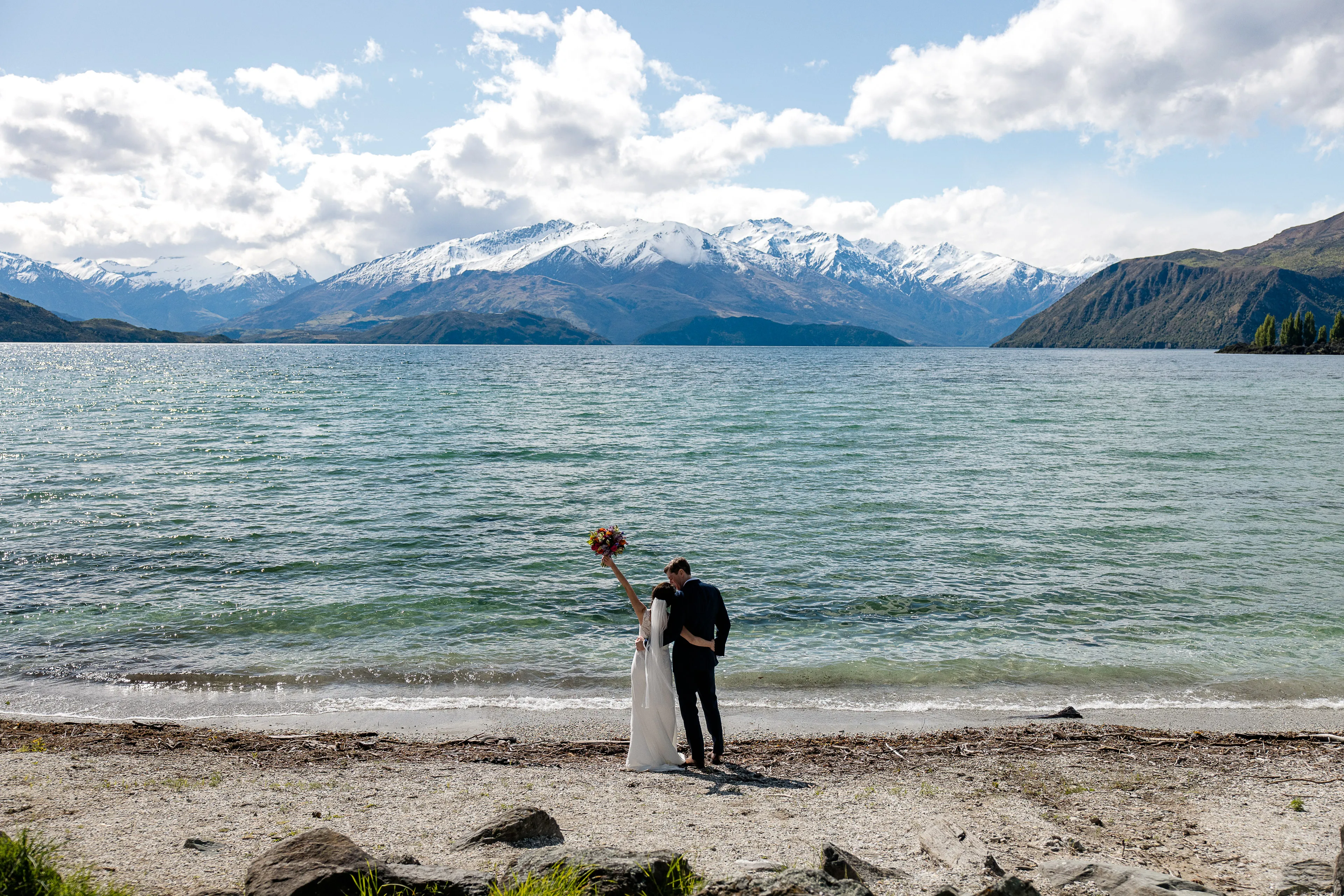 A bride raises her bouquet aloft beside her groom on the shore of Lake Wānaka, the Southern Alps across the lake