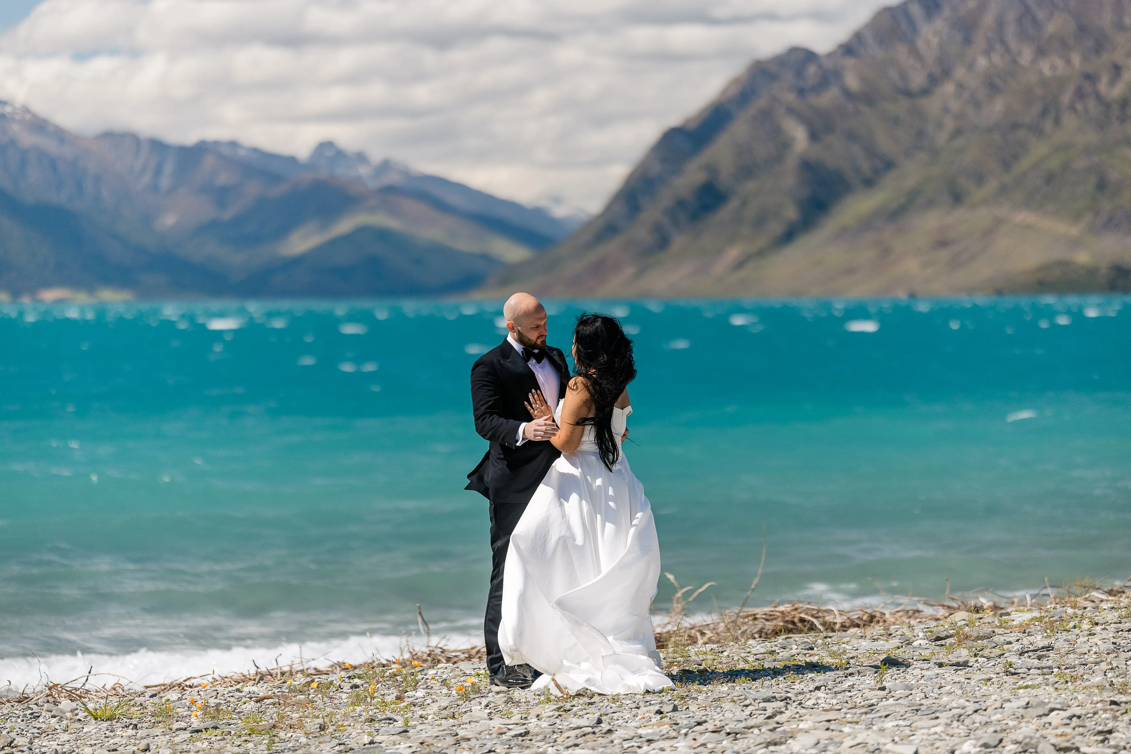 A bride and groom embrace on the pebble shore of Lake Wānaka, vivid turquoise water and mountains behind them