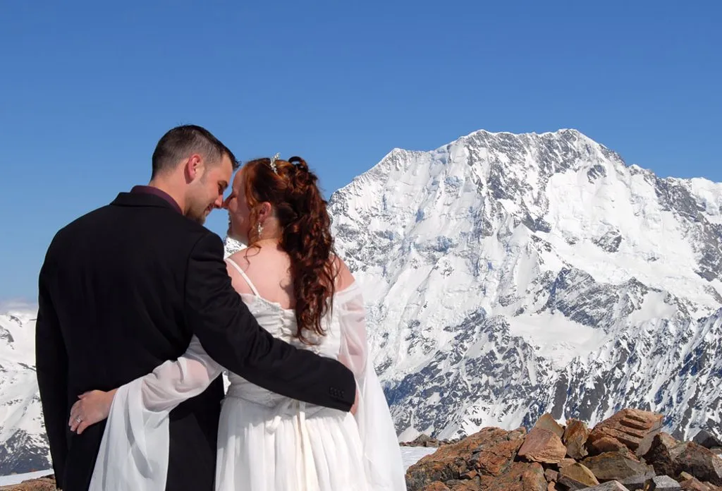 A bride and groom about to kiss on Liebig Dome, a snow-capped peak of Aoraki / Mt Cook behind them