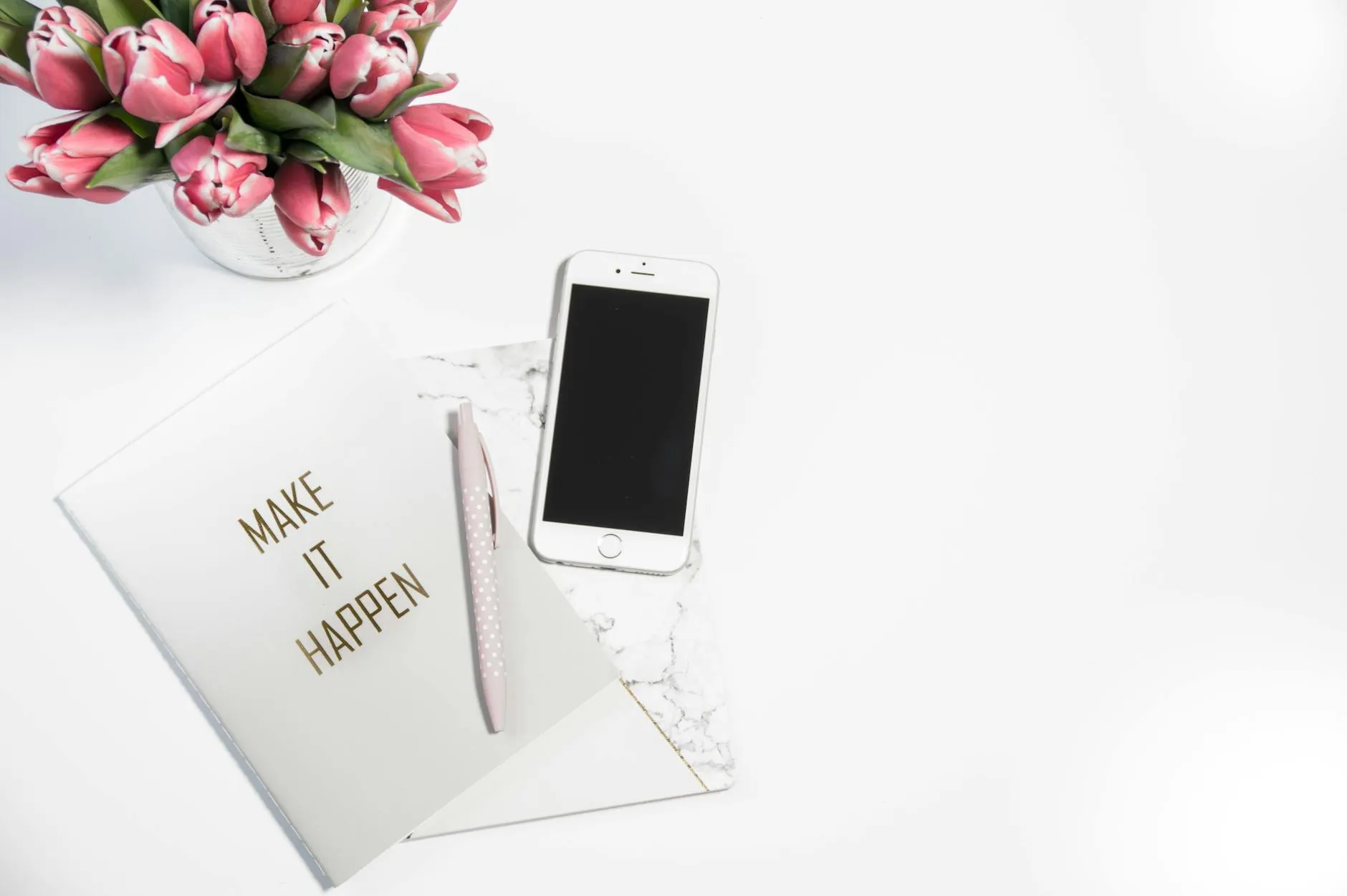 Desk flat lay with pink tulips, a phone and a notebook reading Make It Happen