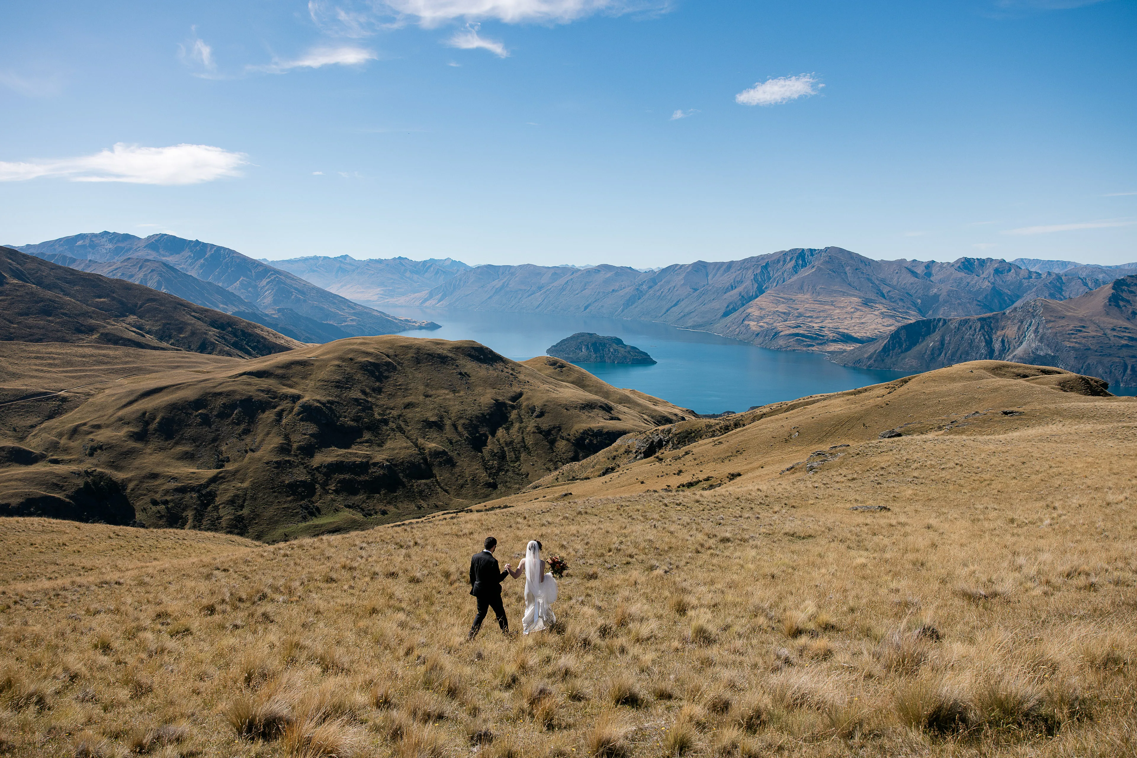 A bride and groom walking through golden tussock toward the lookout over Lake Wānaka