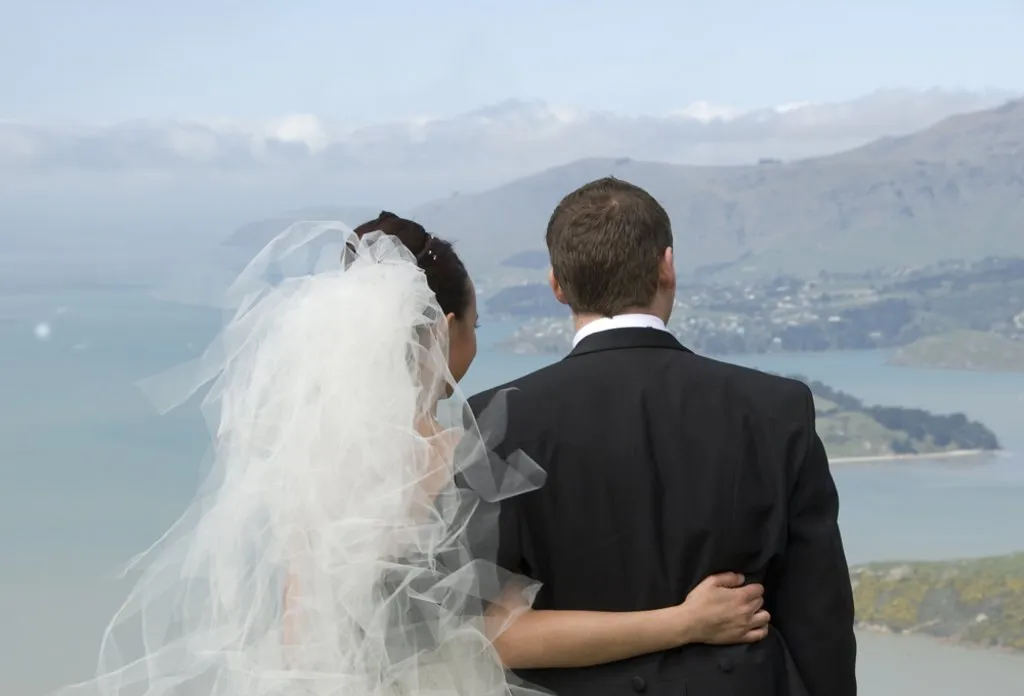 Bride and groom standing arm in arm on a hilltop looking out over a New Zealand lake