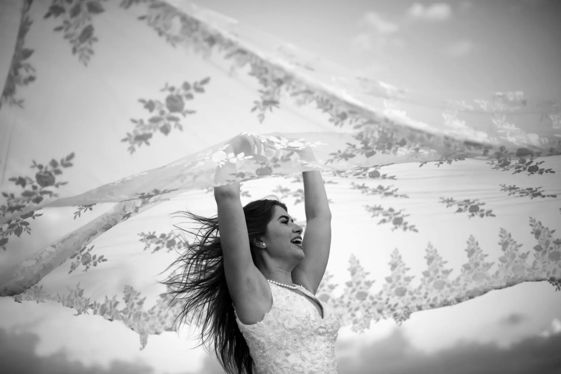 Bride laughing with her arms raised beneath a floral lace veil, black and white