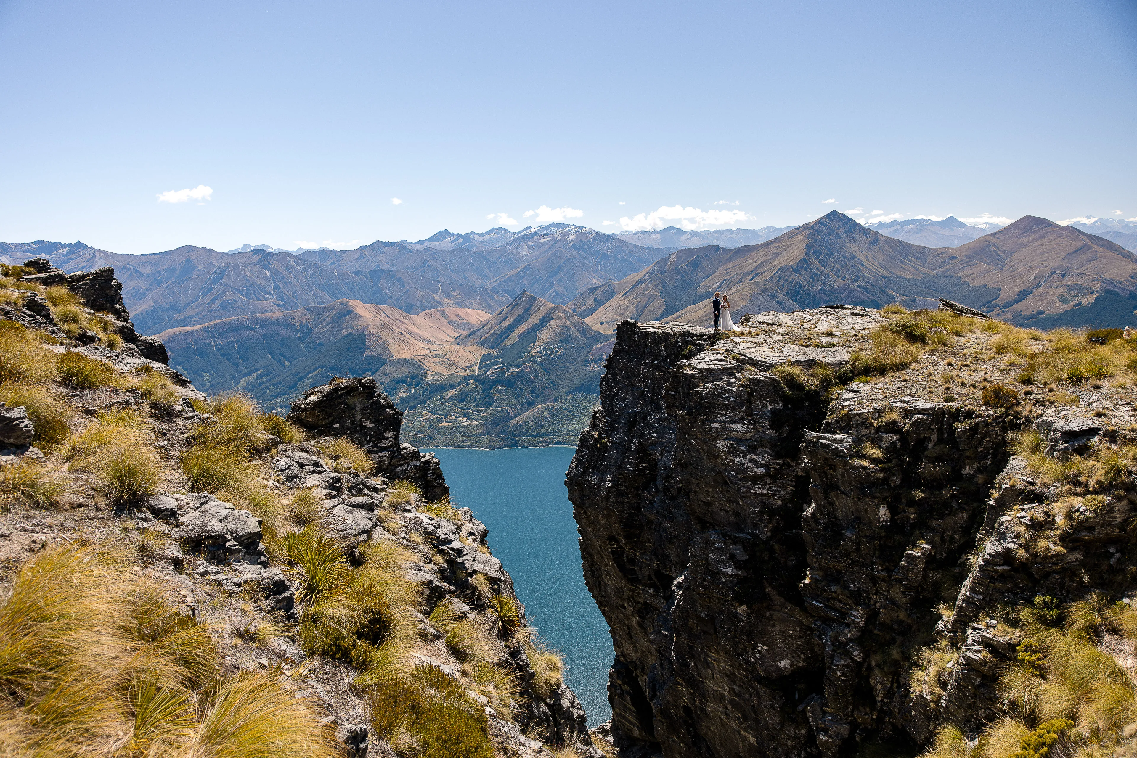 A bride and groom on the dramatic clifftop of The Ledge, Cecil Peak, overlooking Lake Wakatipu