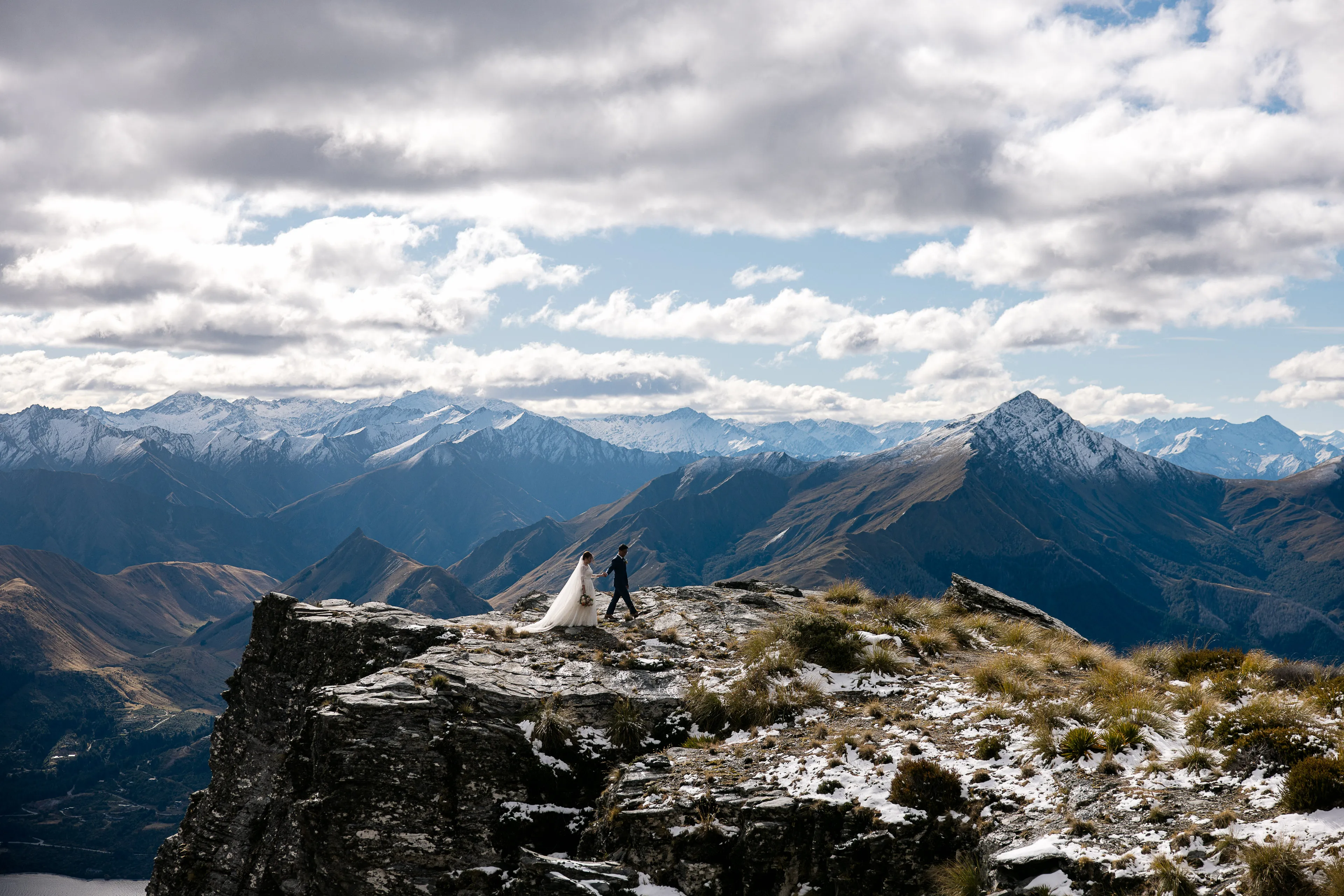 A bride and groom walking along the snow-dusted Ledge on Cecil Peak, panoramic Southern Alps beyond