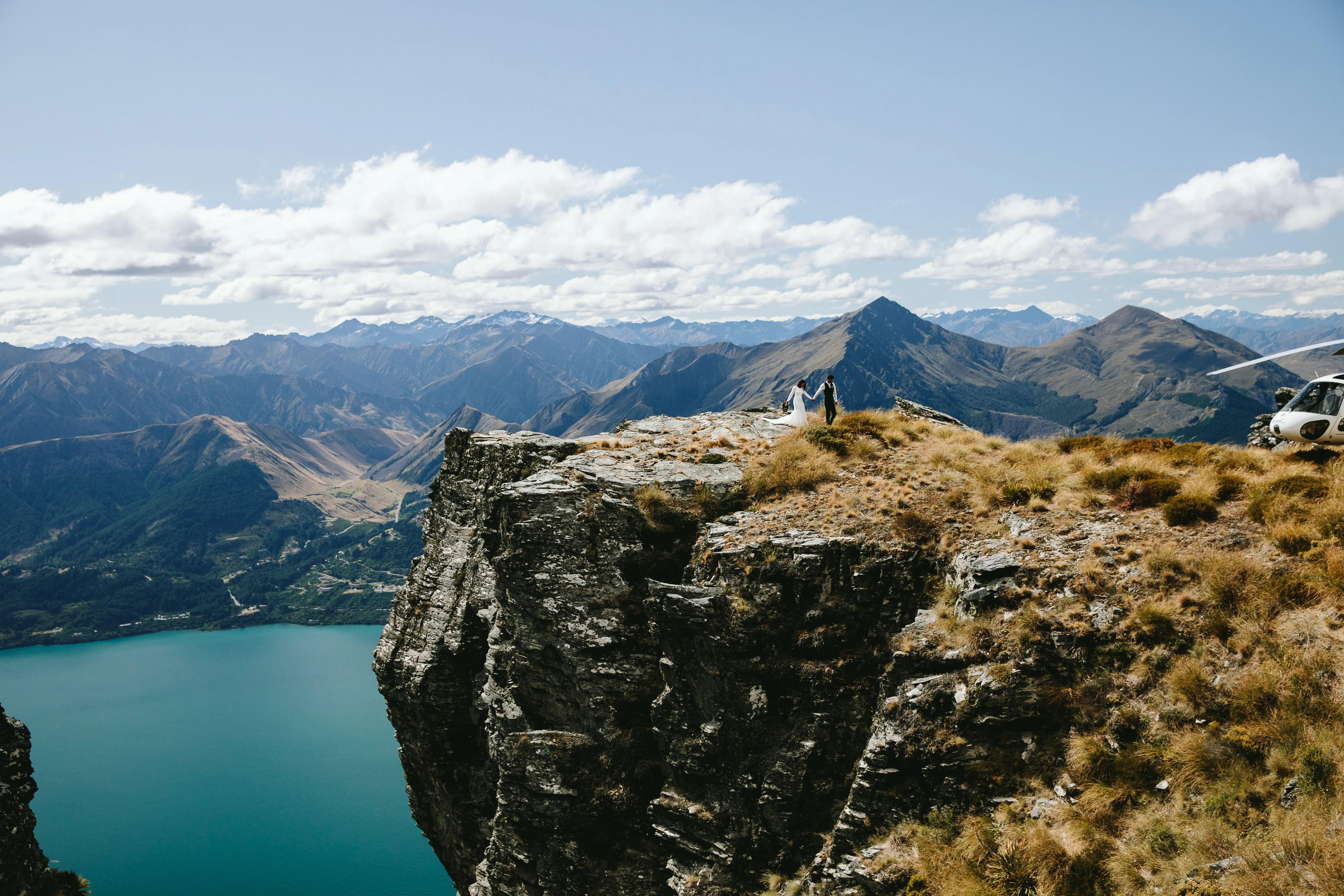 A bride and groom on The Ledge at Cecil Peak beside the helicopter, high above Lake Wakatipu near Queenstown