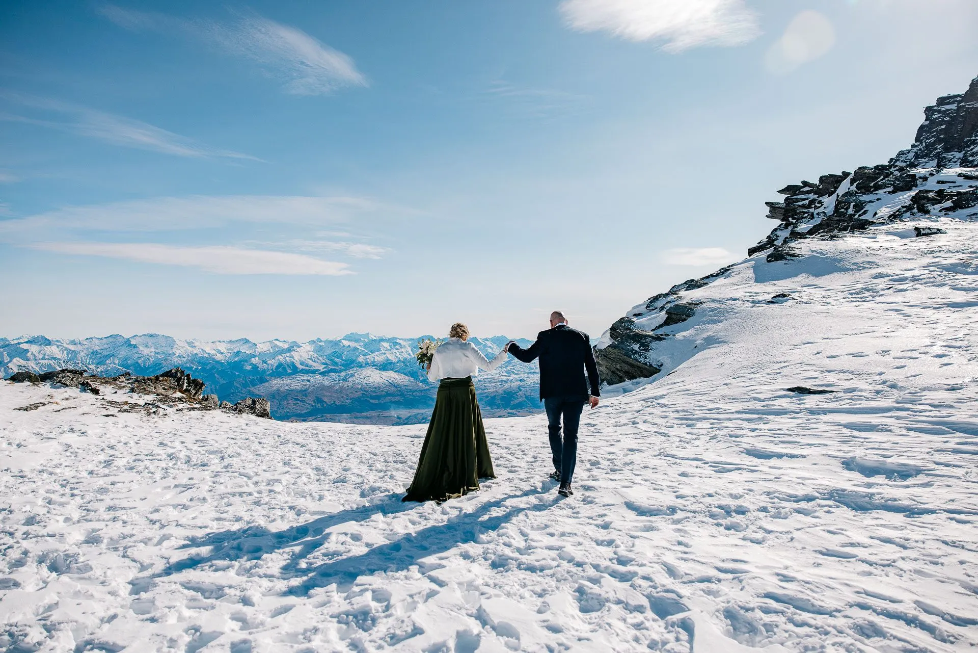 A bride and groom walking hand in hand across the snow on The Remarkables above Queenstown
