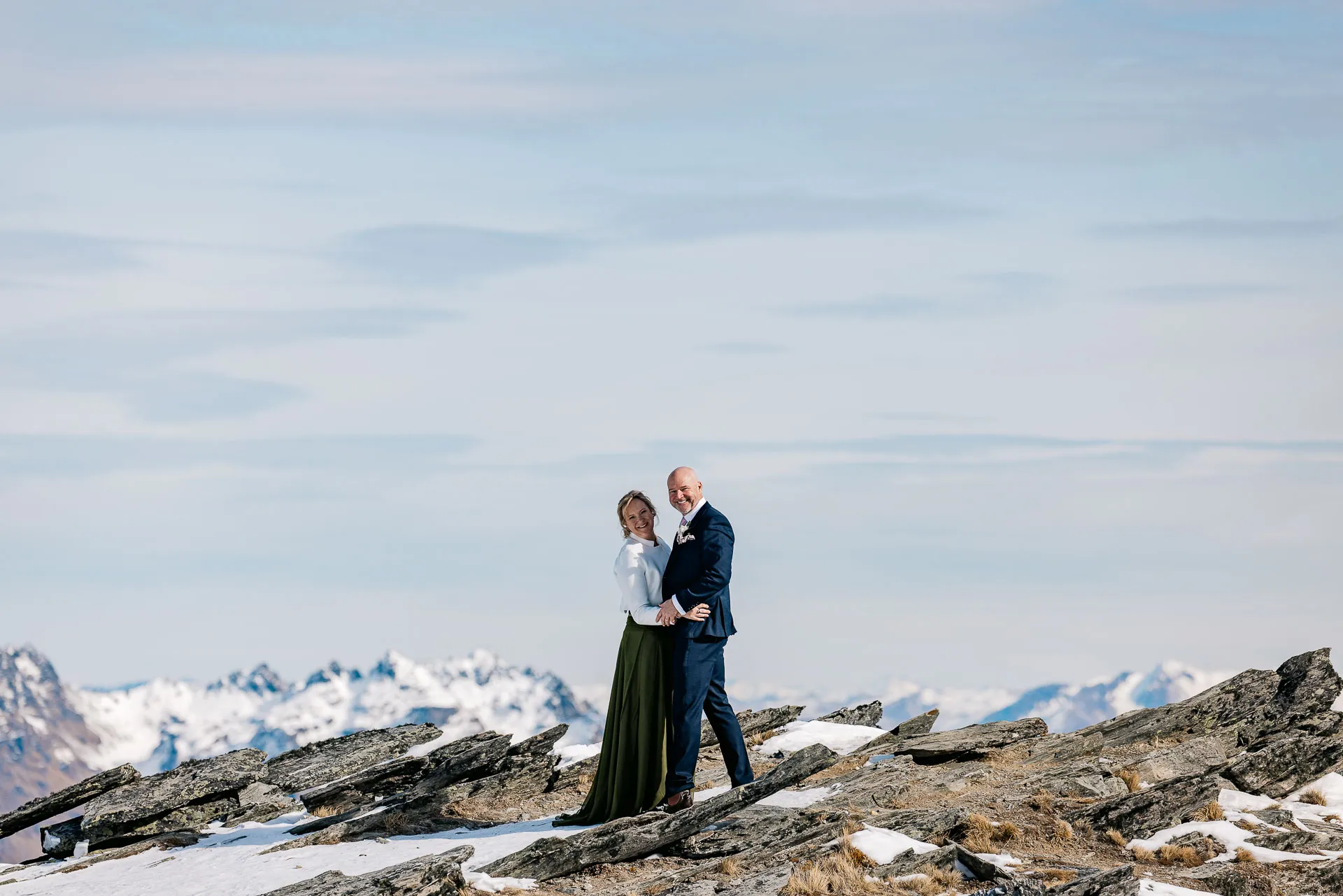 A bride and groom on a rocky ridge of The Remarkables, snow-capped ranges beyond