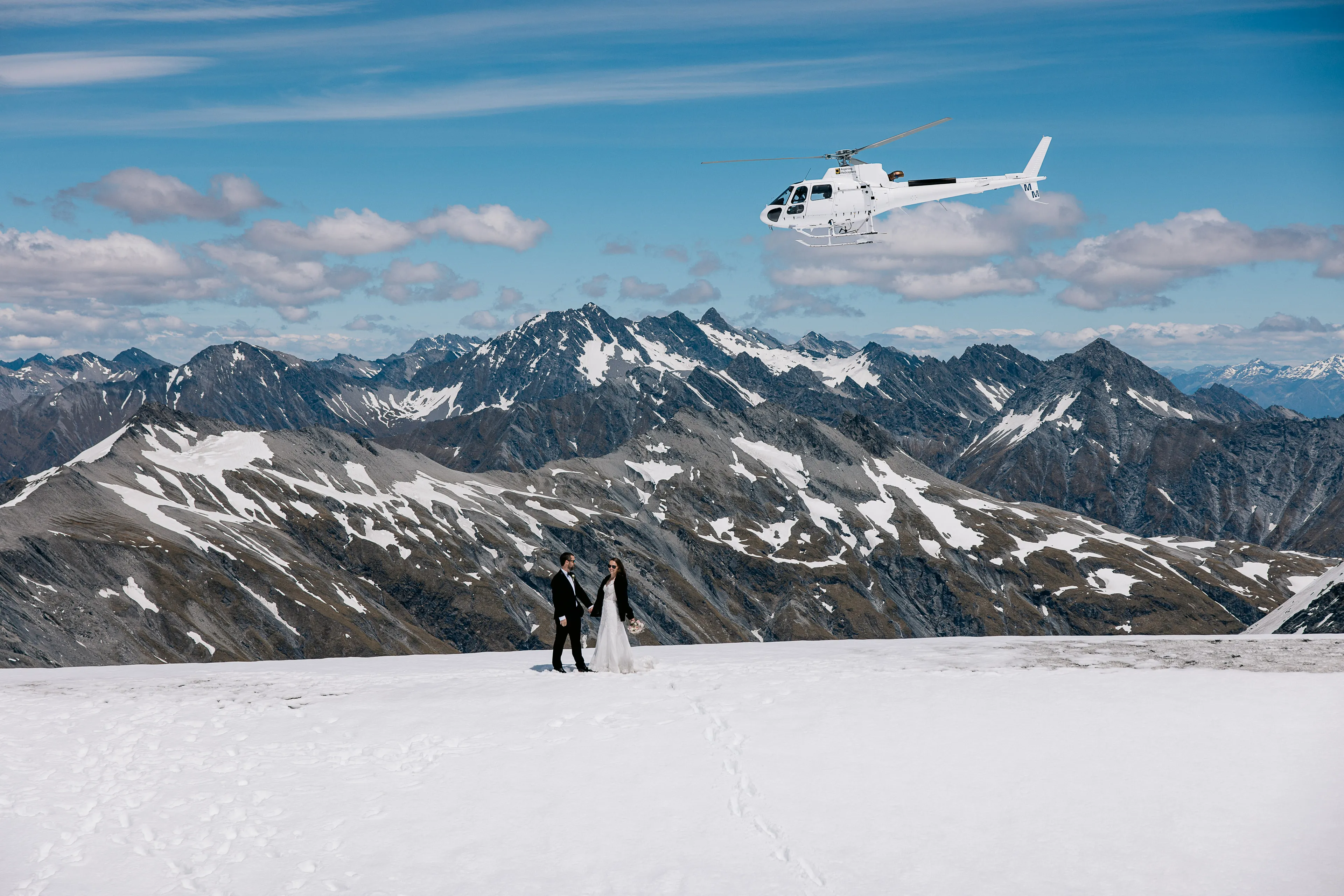 A bride and groom standing on Tyndall Glacier with a helicopter above and the snow-capped Southern Alps behind