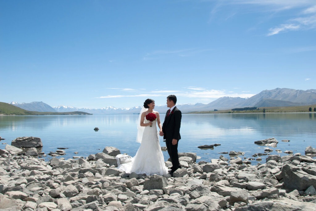 Bride and groom standing by the turquoise waters of Lake Tekapo with mountain views behind them – New Zealand Dream Weddings